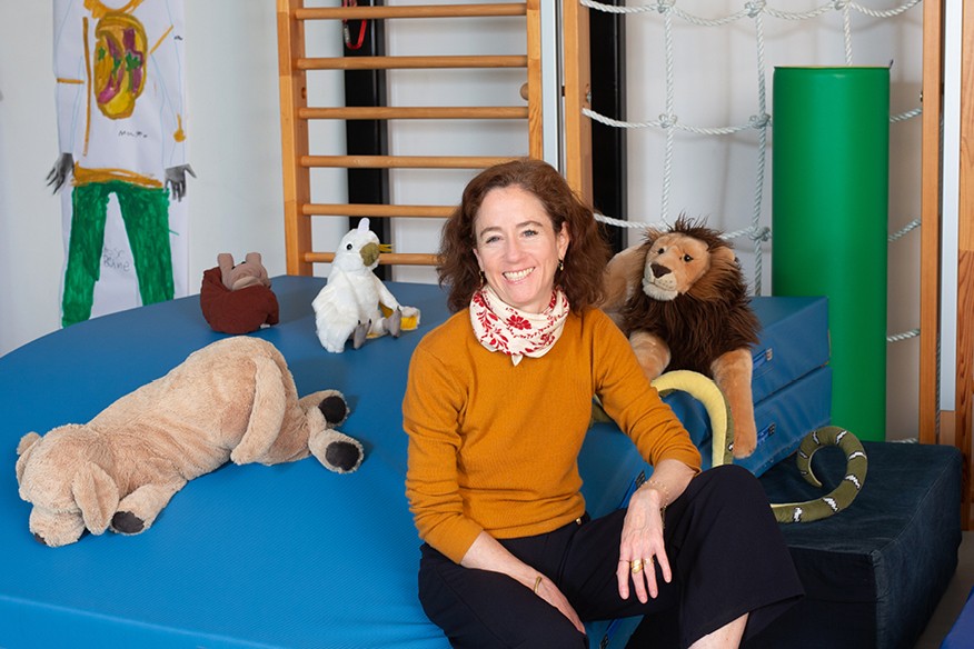 Ariane Macchi in orangem Pullover auf blauer Matte sitzend in Raum für Psychomotorik.