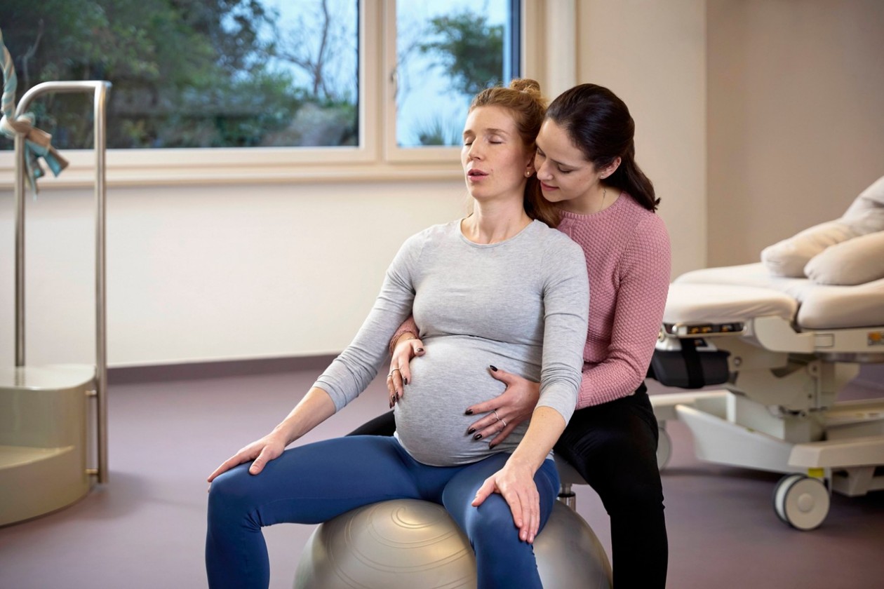 Das Bild zeigt eine schwangere Frau auf einem Sitzball, die von einer Hebamme gestützt wird. 