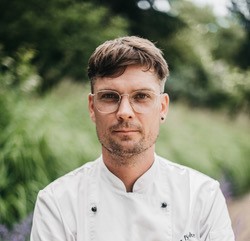 Andi Handke in weisser Kochjacke. Er trägt eine Brille. Der Hintergrund ist weichgezeichnet und grün. 