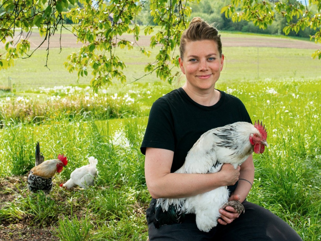 Man sieht Anna Pearson, sie hält ein weisses Huhn im Arm, im Hintergrund eine grüne Wiese und zwei weitere Hühner.