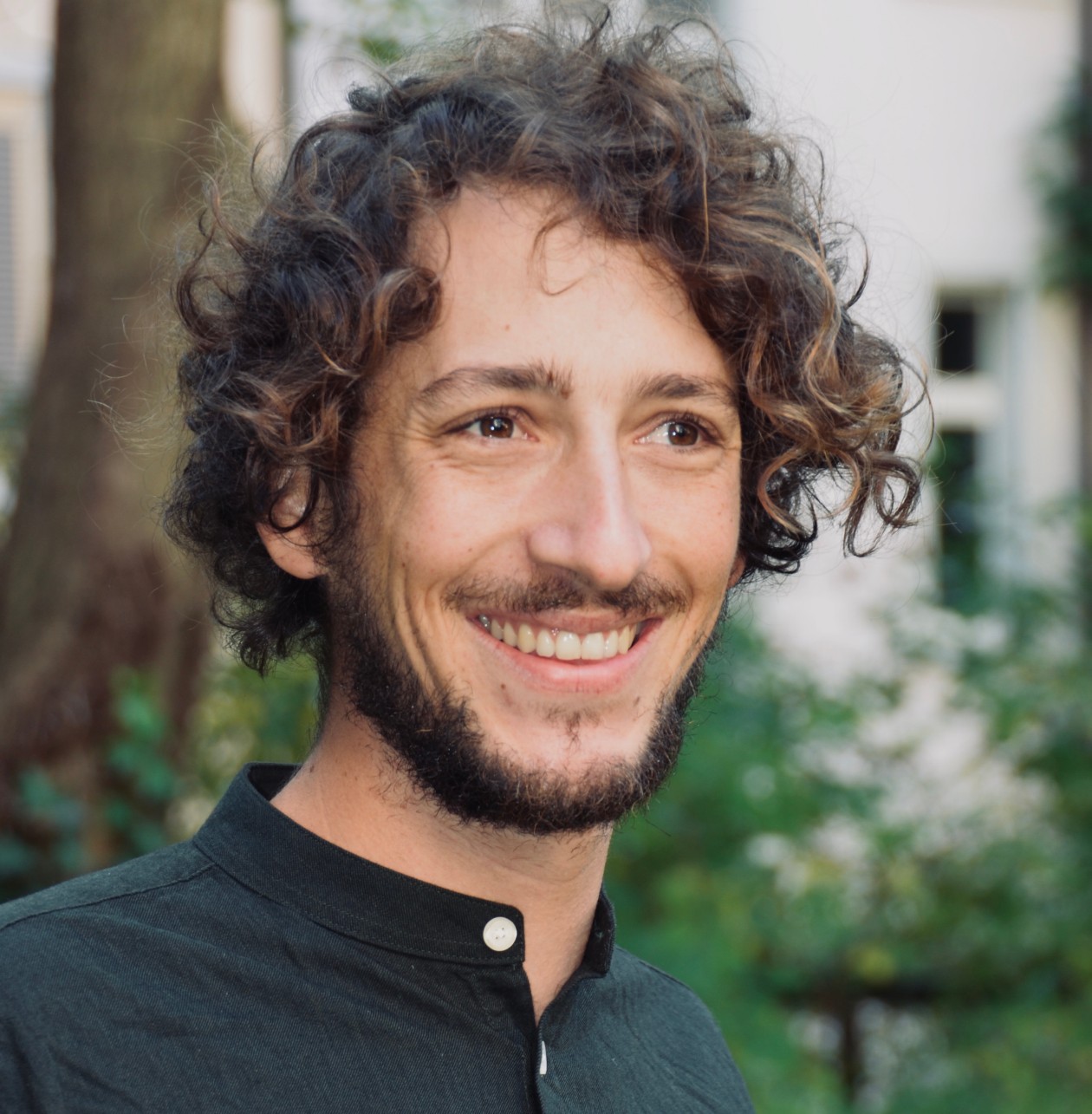 Lukas Domeisen trägt ein schwarzes Hemd, er blick in die Kamera und lächelt. Im Hintergrund grüne Sträucher.