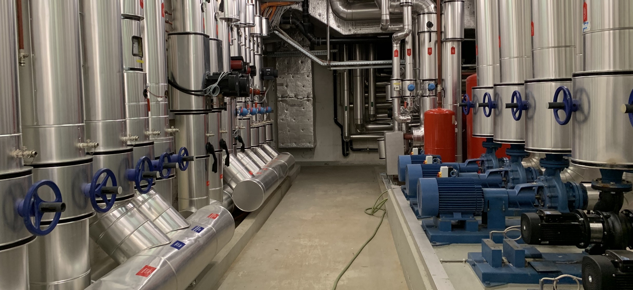 Ansicht einer Heizungsverteileranlage, links und rechts dicke Leitungen, in der Mitte ein Gang