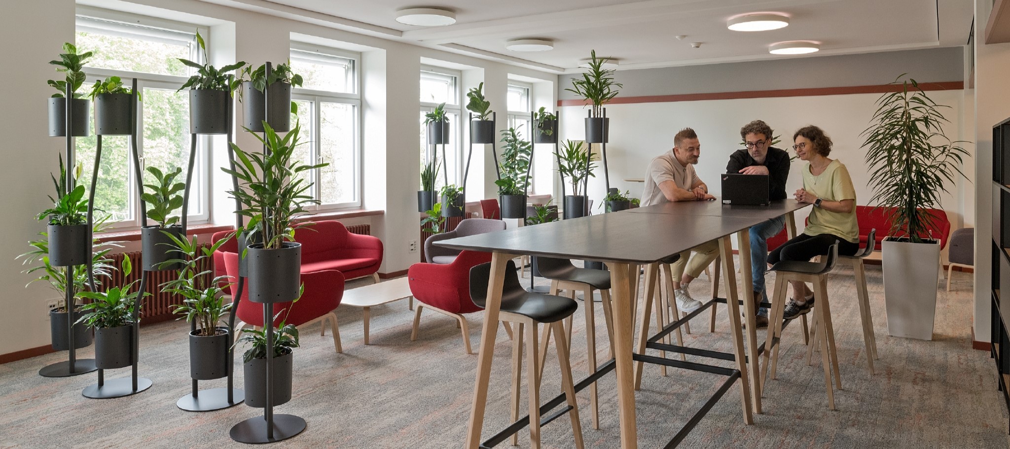 Aufenthaltsraum mit roten Sofas, Pflanzen, zwei Männer und eine Frau sitzen an einem Hochtisch und blicken auf den Bildschirm eines Laptops