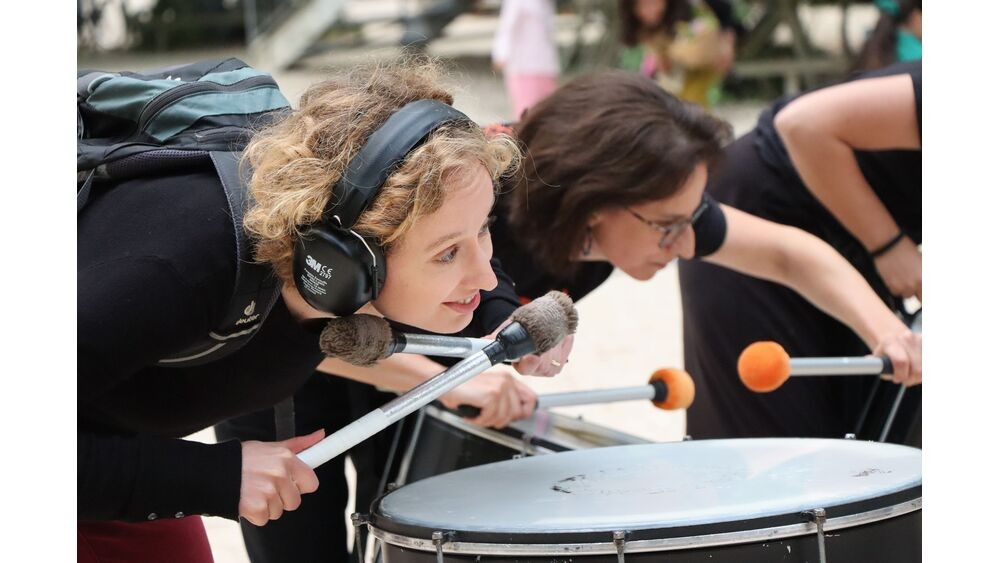 Zwei Musikerinnen stehen vor grossen Trommeln und halten während des Spiels ihr Ohr nahe zum Instrument.  