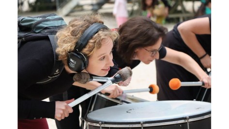 Zwei Musikerinnen stehen vor grossen Trommeln und halten während des Spiels ihr Ohr nahe zum Instrument.  
