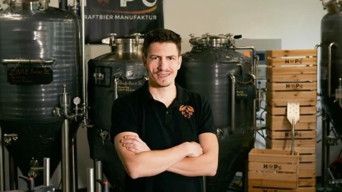 Ein Brauer steht im schwarzen T-Shirt zwischen Harassen und Biertanks.