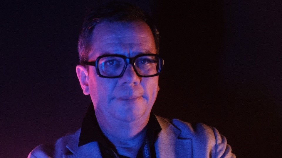 Alex Bücheli mit grosser Hornbrille steht im Dunkel eines Clubs und wird von violettem Licht angeleuchtet. 