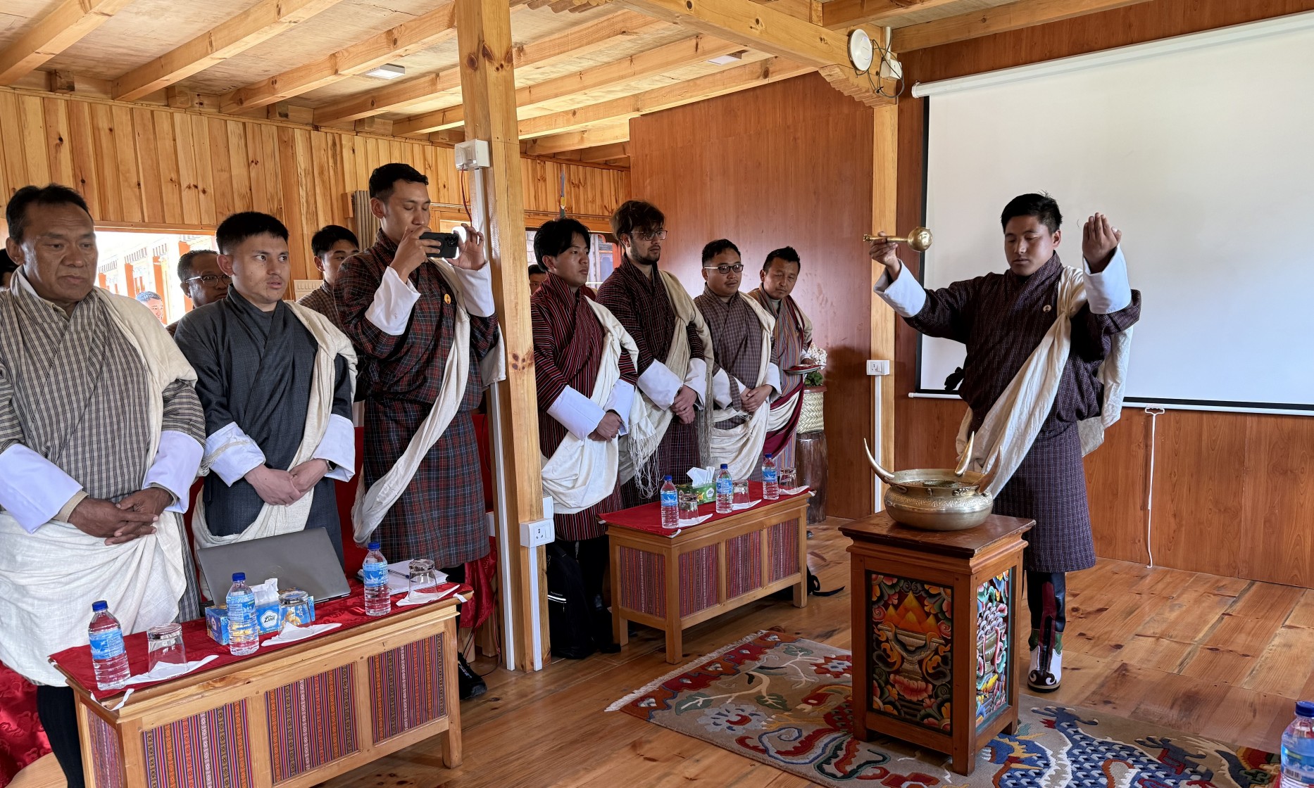 Eröffnungszeremonie der Workshops: In einem mit Holz ausgekleideten Raum stehen Männer aus Bhutan, alle mit einer Schärpe um den Oberkörper. Vorne, vor einer Art kleiner Altar, steht ein Mann mit gehobenen Armen. 
