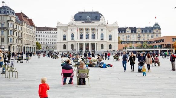 Sechseläutenplatz mit Sicht auf das Opernhaus. Auf dem Platz sitzen, stehen und gehen viele Leute.