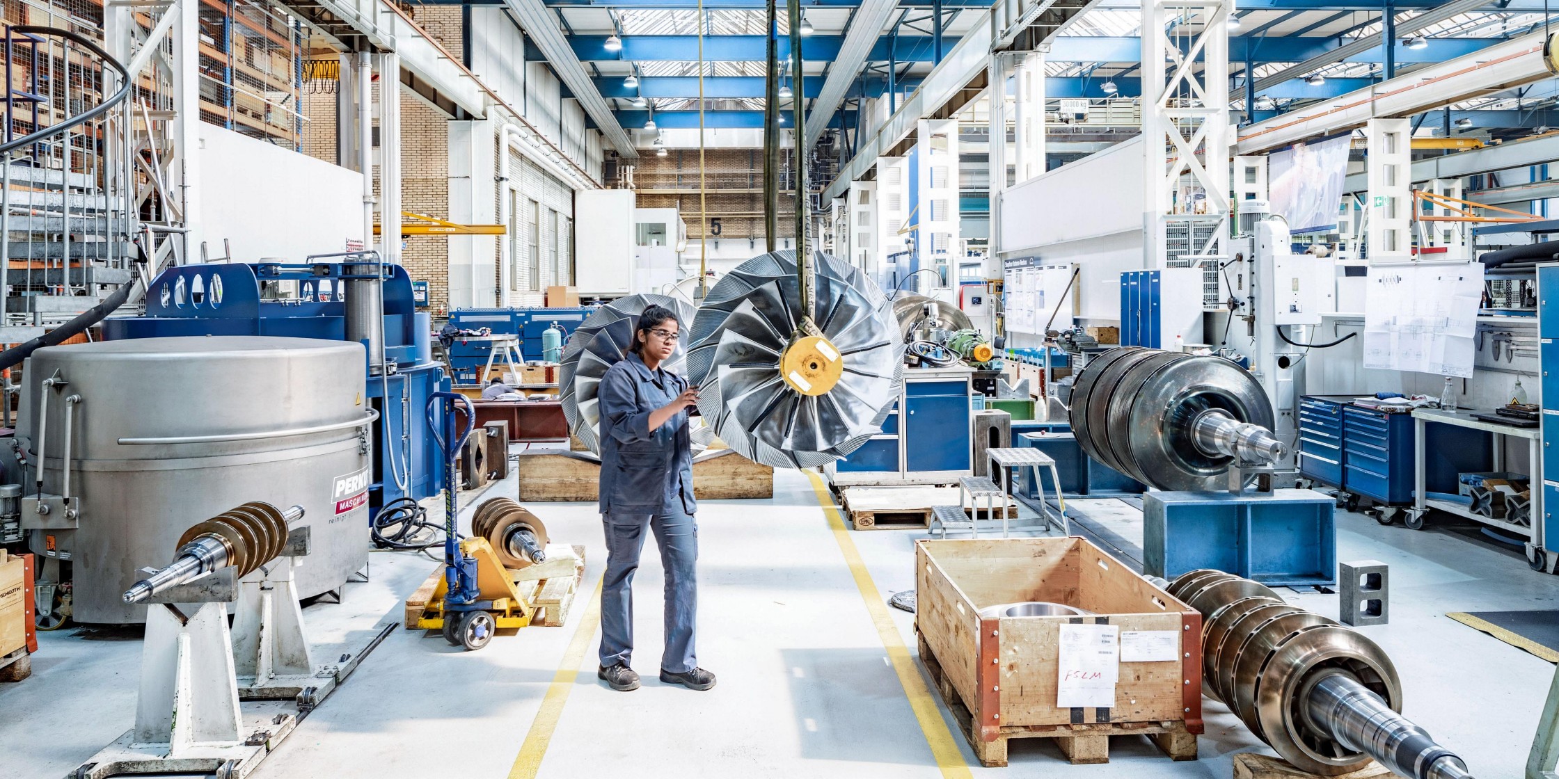 In einer Industriehalle steht eine junge Frau im blauen Overall und arbeitet an einem Rad, das von der Decke hängt. 