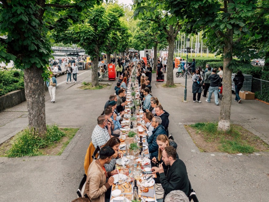 Menschen sitzen unter einem endlos langen Tisch unter Bäumen und essen. 