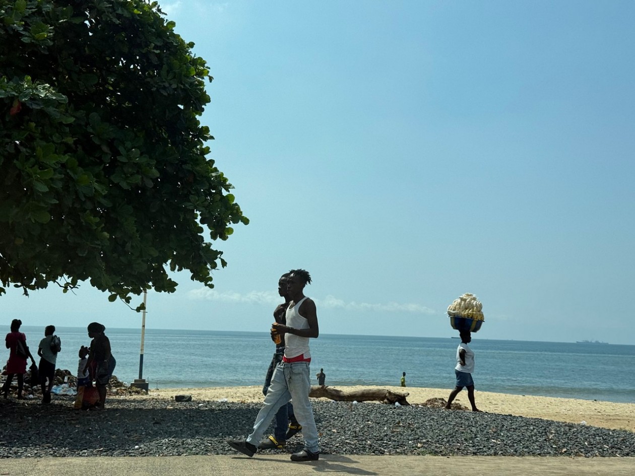 An der Strandpromenande laufen junge Leute in Jeans und eine Frau mit einem Strunk Bananen auf dem Kopf balancierend vorbei.  