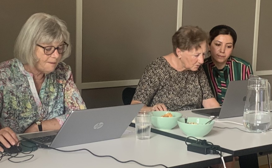 Zwei ältere Frauen sitzen am Computer. Eine jüngere Frau unterstützt sie. 