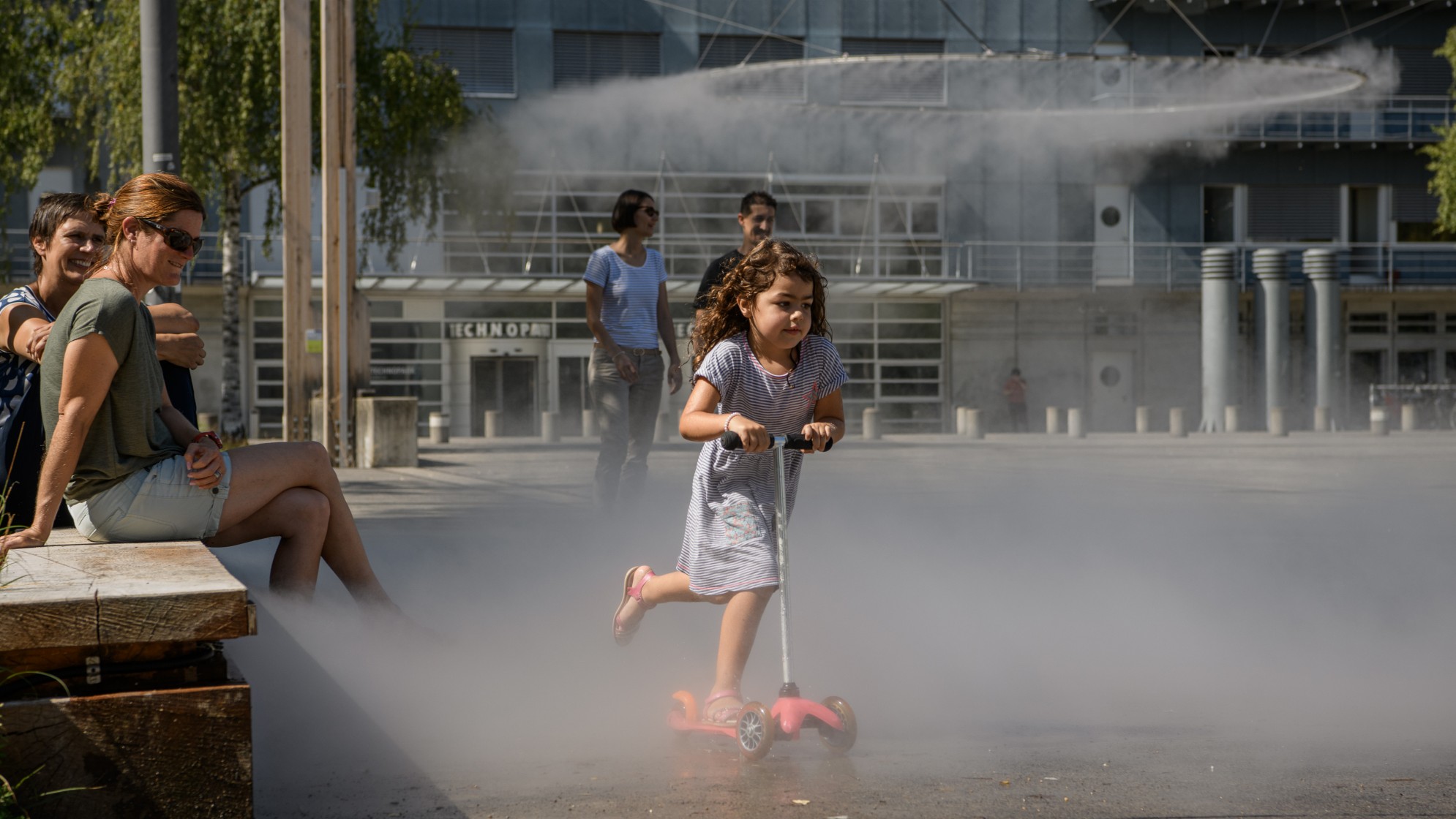 Ein kleines Mädchen fährt mit dem Trottinett unter dem Sprühnebel der Wolke hindurch.