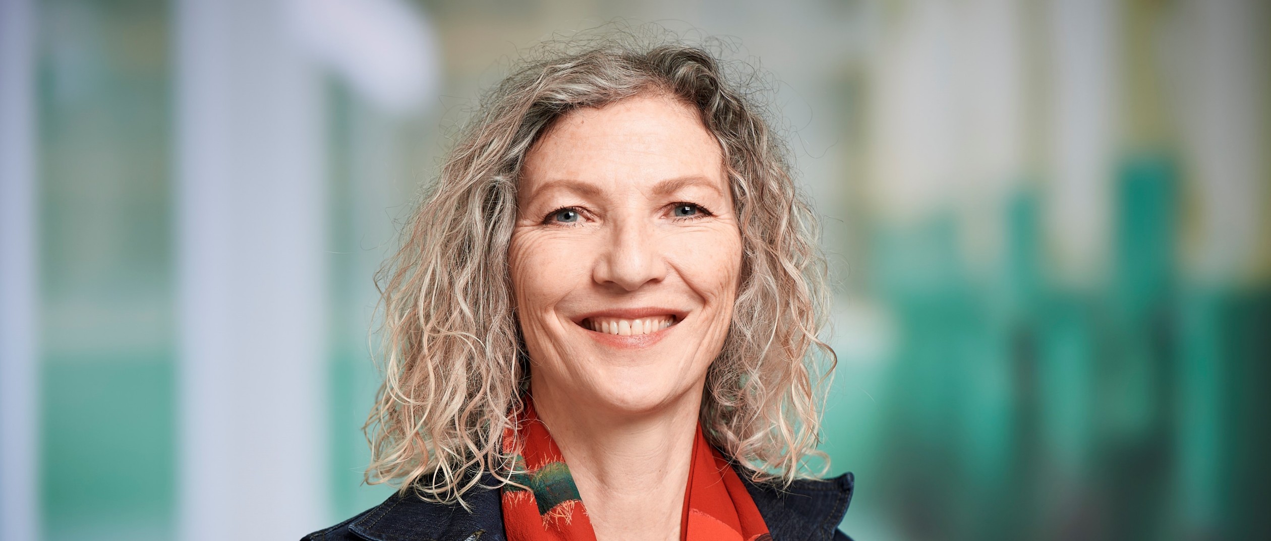 Silva Rouss, eine Frau mit  blonden Locken und buntem Foulard, lacht in die Kamera.