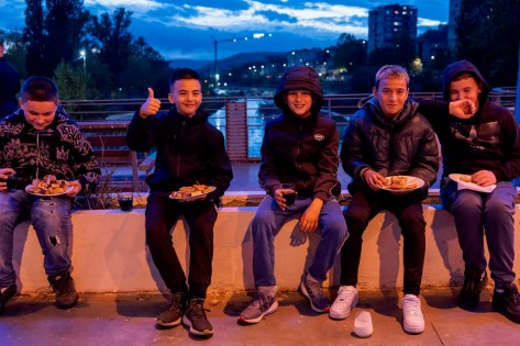 Fünf Knaben sitzen beim Eindunkeln nebeneinander auf einer Brücke, haben gefüllte Teller in der Hand und lachen für den Fotografen. 