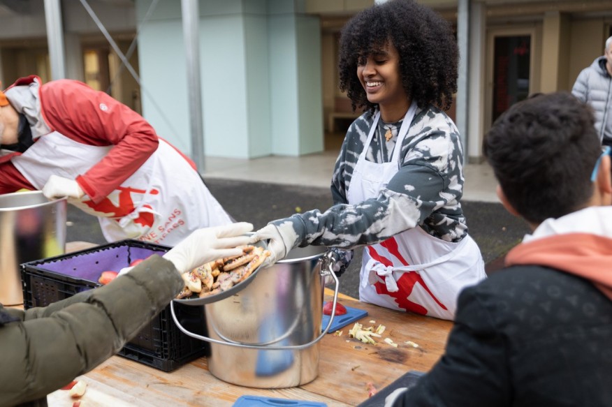 Eine junge Frau mit dunklen Locken steht hinter einem grossen Topf und reicht einer ausgestreckten Hand einen teller Essen.