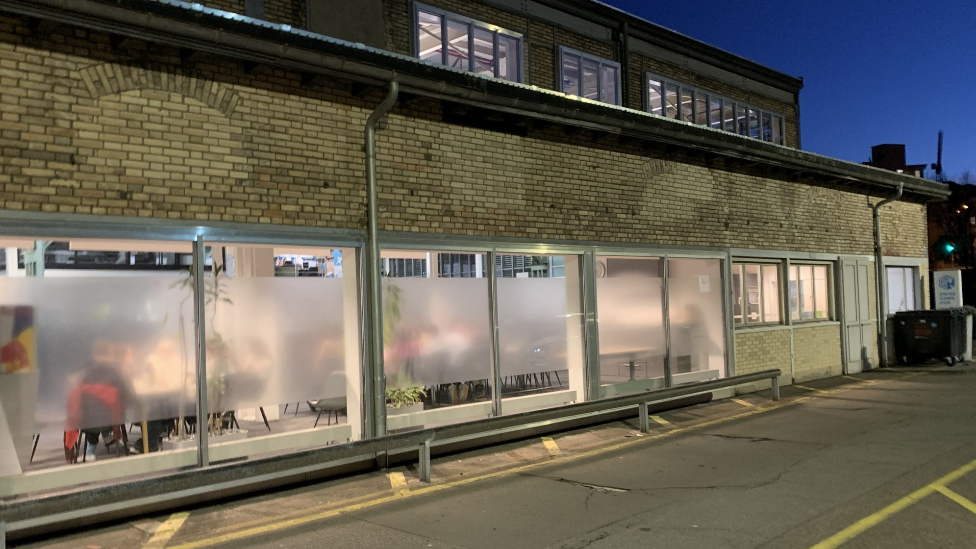 Die Backsteinfassade der früheren Maschinenfabrik. Drinnen brennt Licht, man erkennt schemenhaft Leute, die an langen Tischen zusammensitzen.