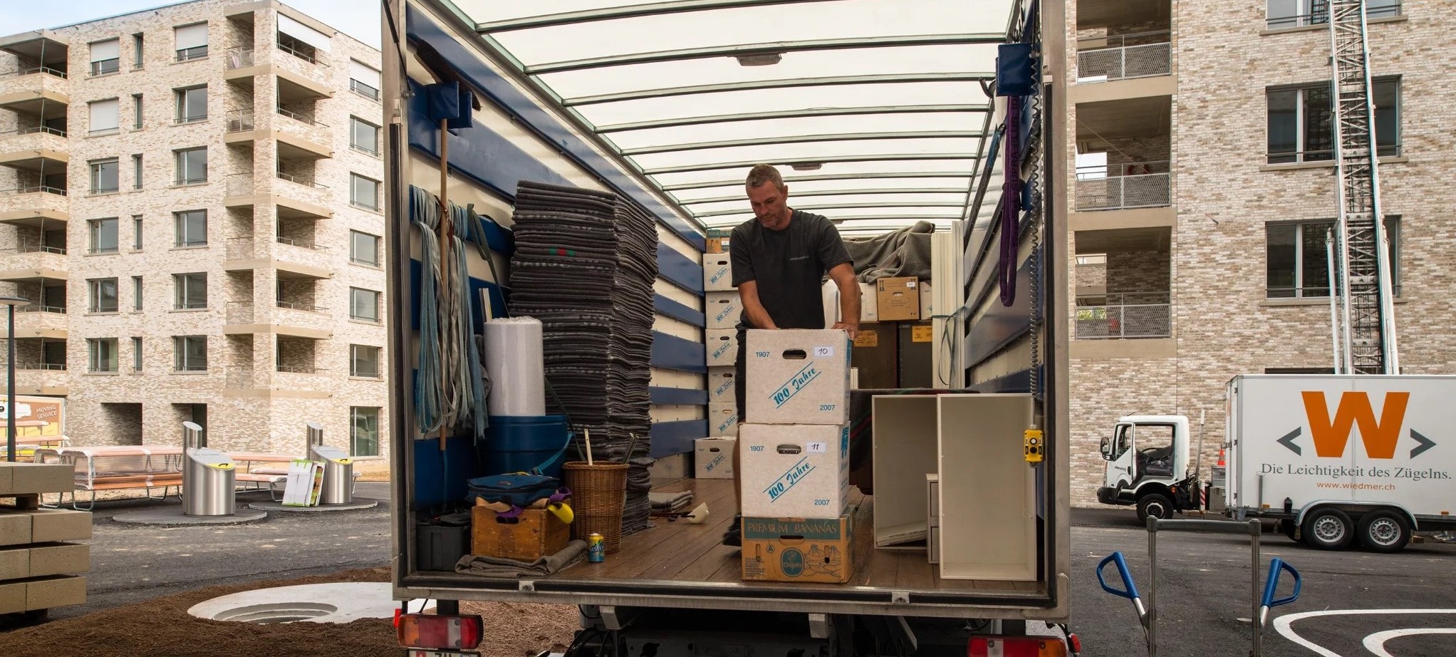 Vor neubauten steht ein offener Zügelwagen, ein Mann lädt Kisten ein.