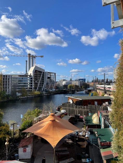 Holzmarkt_1 An einem Fluss stehen auf der linken Seite Hochhäuser und Kräne, auf der rechten kleine, bunte Hütten und Sonnendächer.