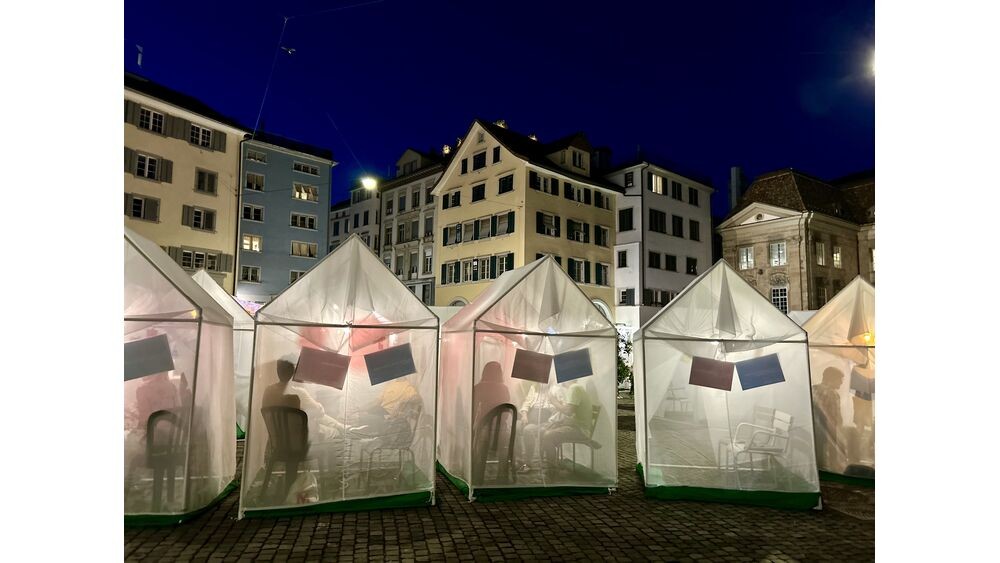 Der Münsterhof bei Nacht: Mehrere kleine durchscheindende Zelte stehen aneinander, dahinter erkennt man schemenhaft sitzende Menschen im Gespräch. 