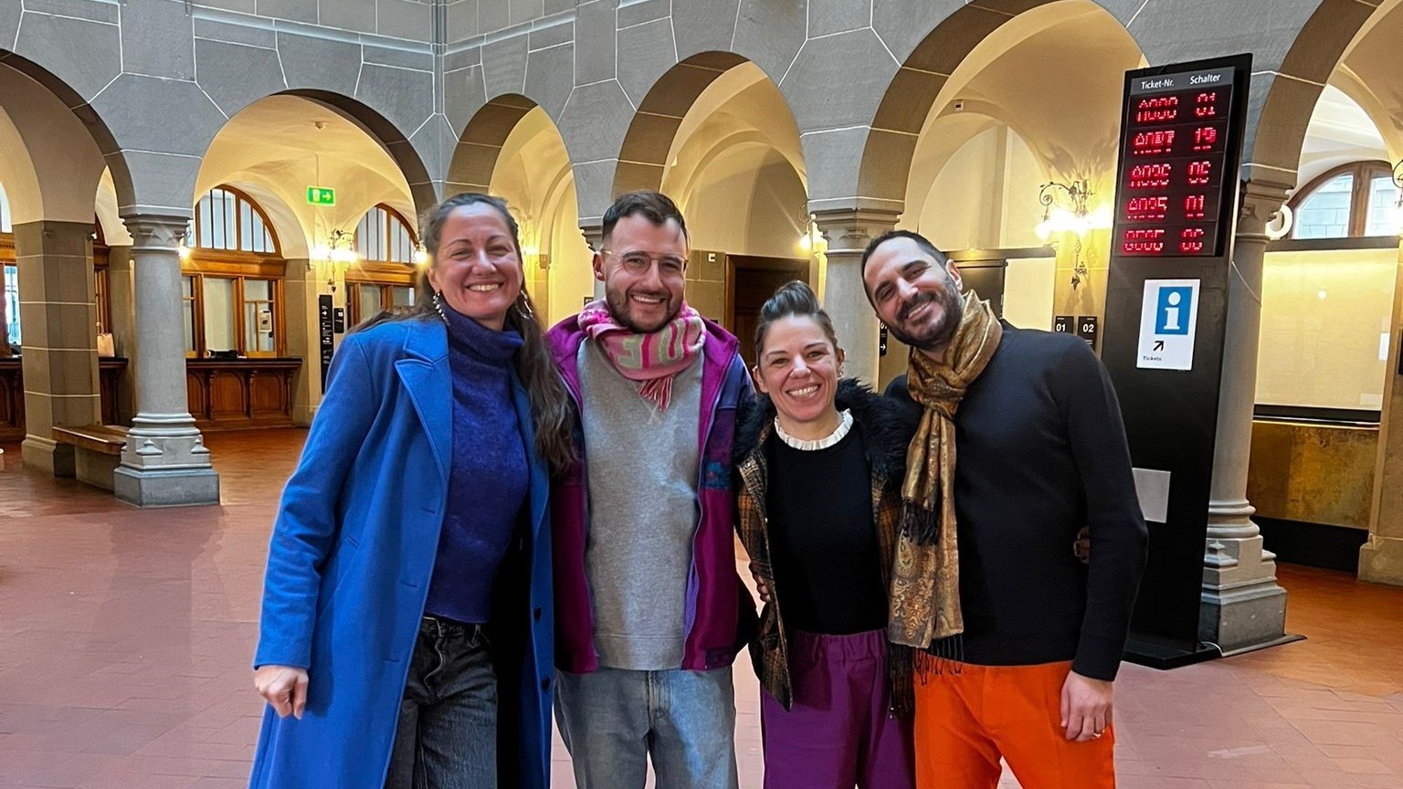 Zwei Frauen und zwei Männer stehen in der Eingangshalle des Stadthauses. Sie haben sich die Arme freundschaftlich über die Schultern gelegt und lachen in die Kamera. 