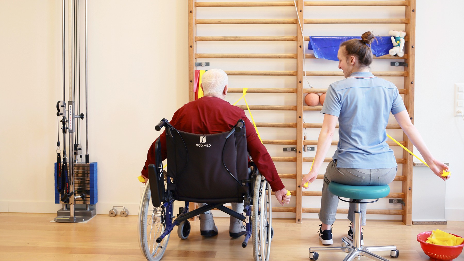 Bewohnerin im Rollstuhl macht vor einer Sprossenwand Übungen mit einer Therapeutin.