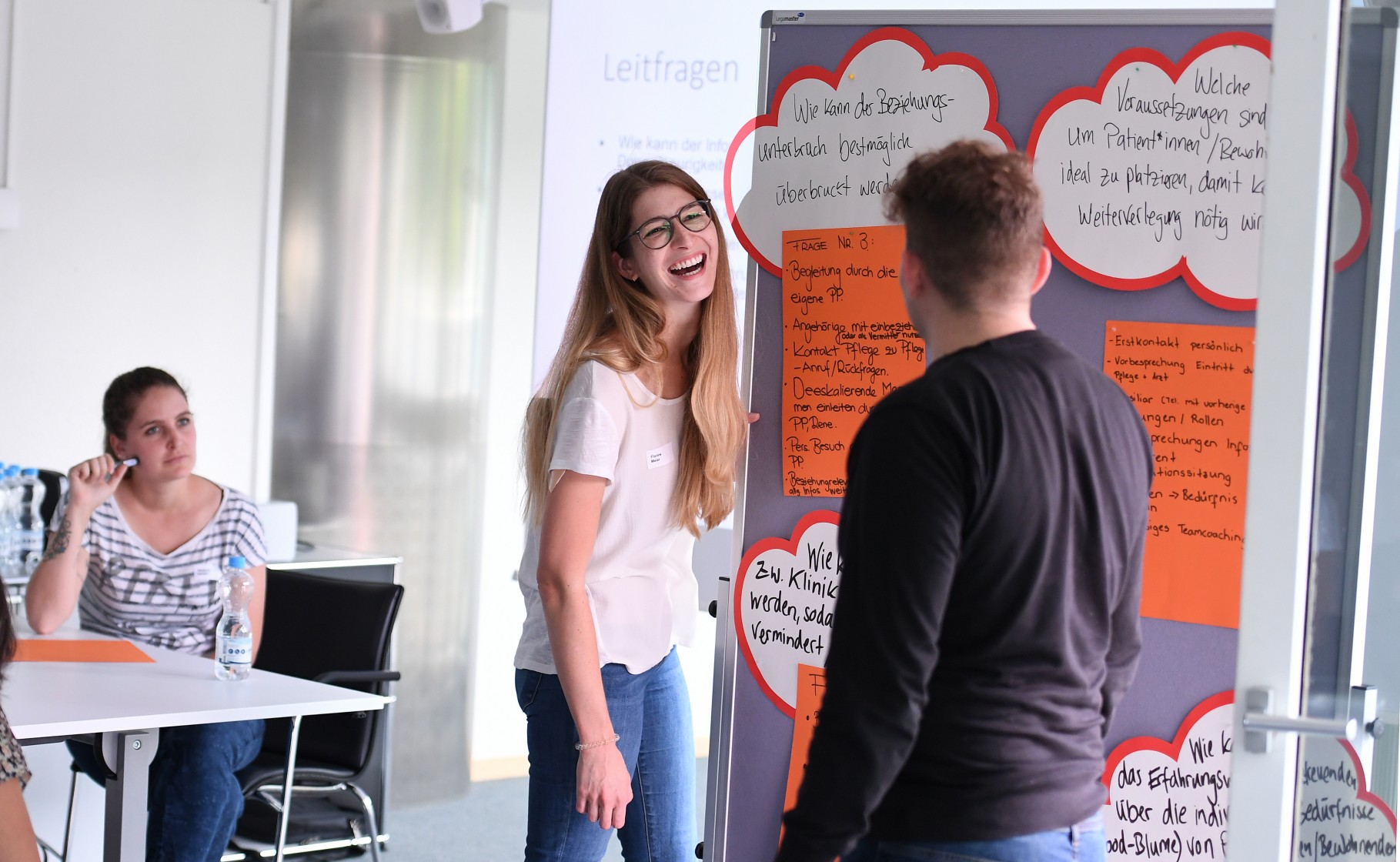Drei Studierende diskutieren eine Gruppenarbeit und kleben ihre Gedanken an eine Pinnwand