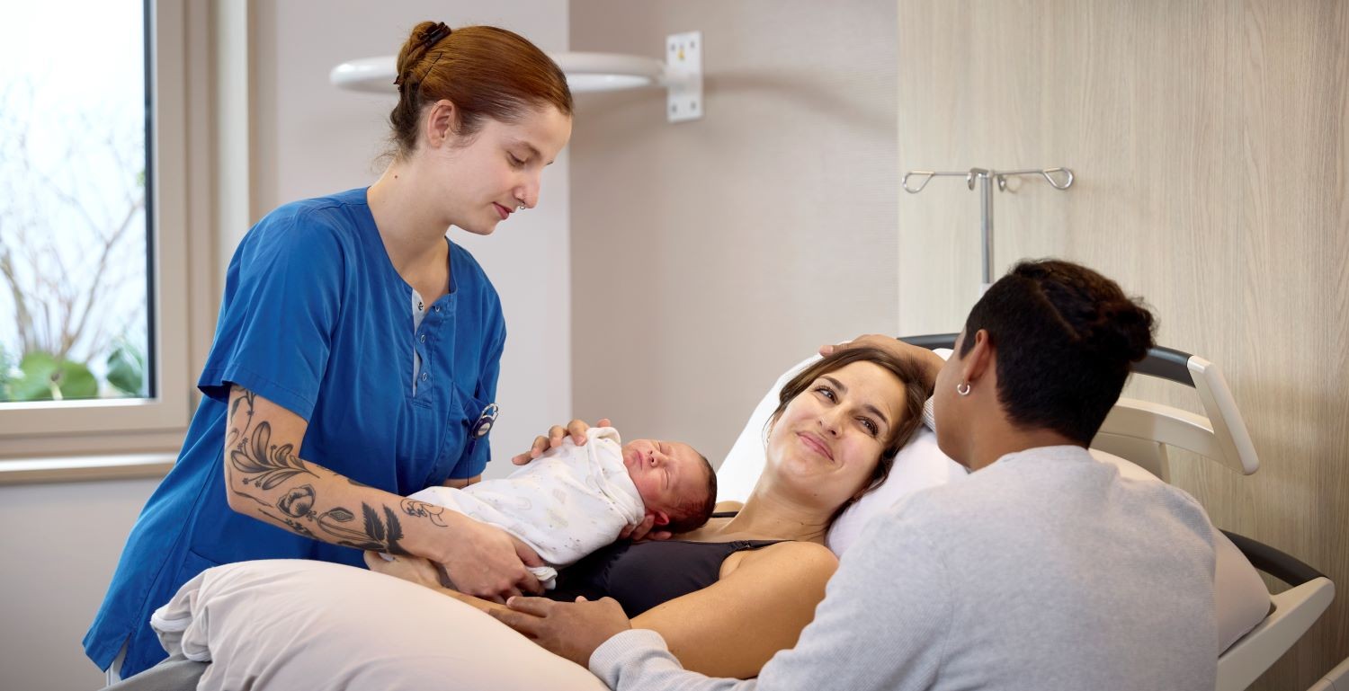 Hebamme überreicht Mutter das Baby und der Vater am Bett