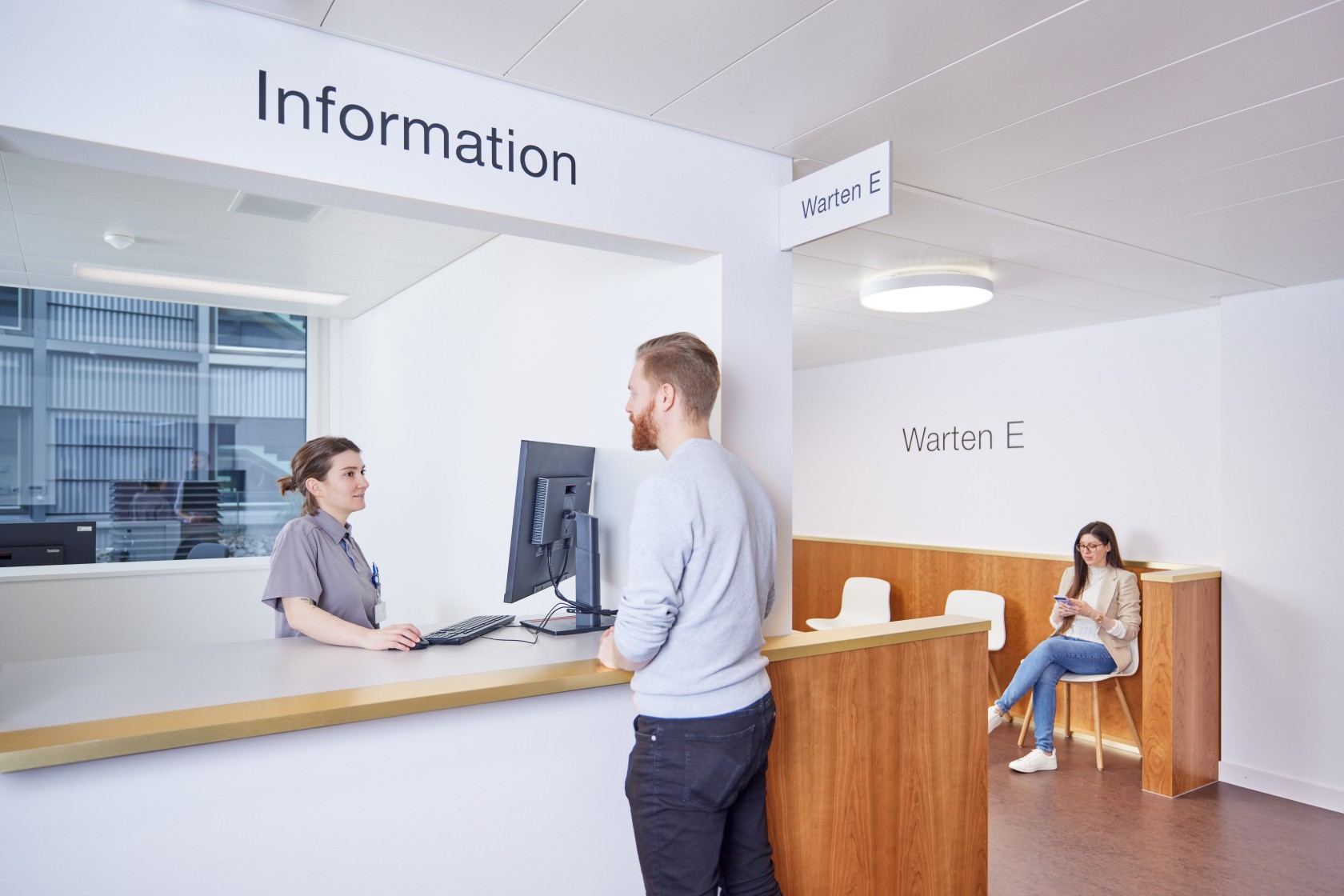 Ein Mann meldet sich bei der Praxis-Assistentin an. Im Wartebereich sitzt eine Patientin.