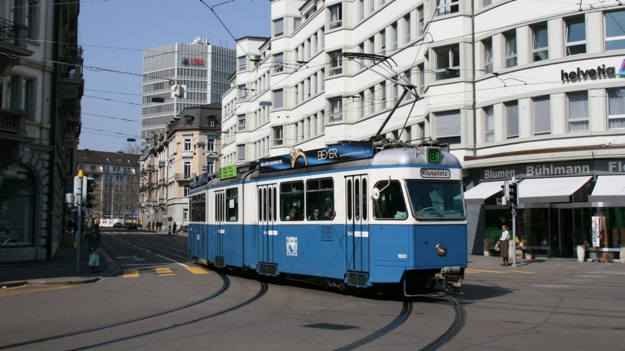 Eine blau-weiße Tram fährt durch eine Strassenkreuzung in einer städtischen Umgebung. Im Hintergrund sind moderne und historische Gebäude zu sehen.