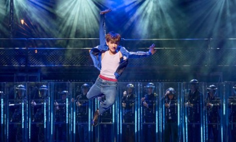 Intranet Teaserbild - Billy Elliot Billy Elliot springt in die Luft, im Hintergrund stehen Polizisten in Uniform.