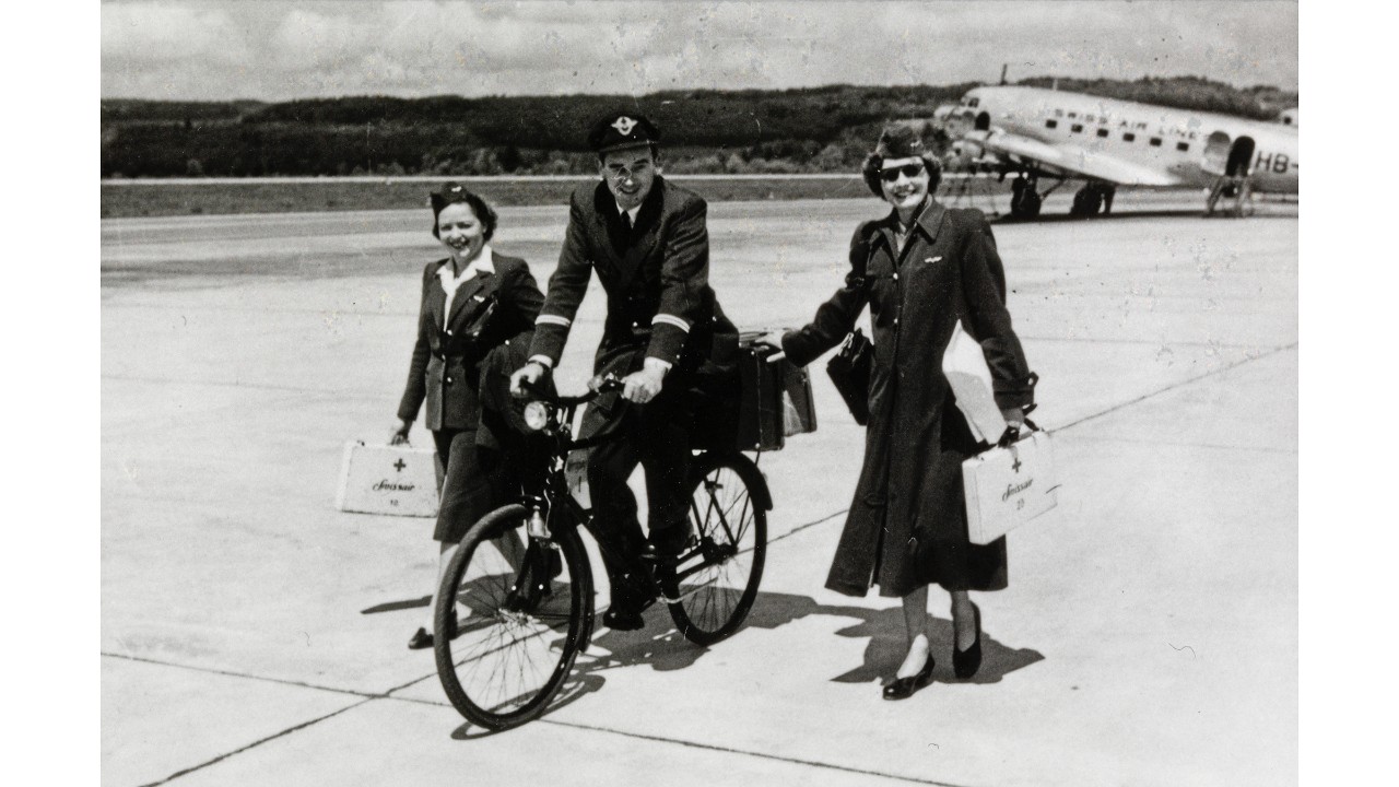 Um 1950, ein Swissair-Pilot auf einem Pistenvelo und in Begleitung von zwei Flugbegleiterinnen am Flughafen Kloten (Quelle: ETH-Bibliothek Zürich, Bildarchiv)