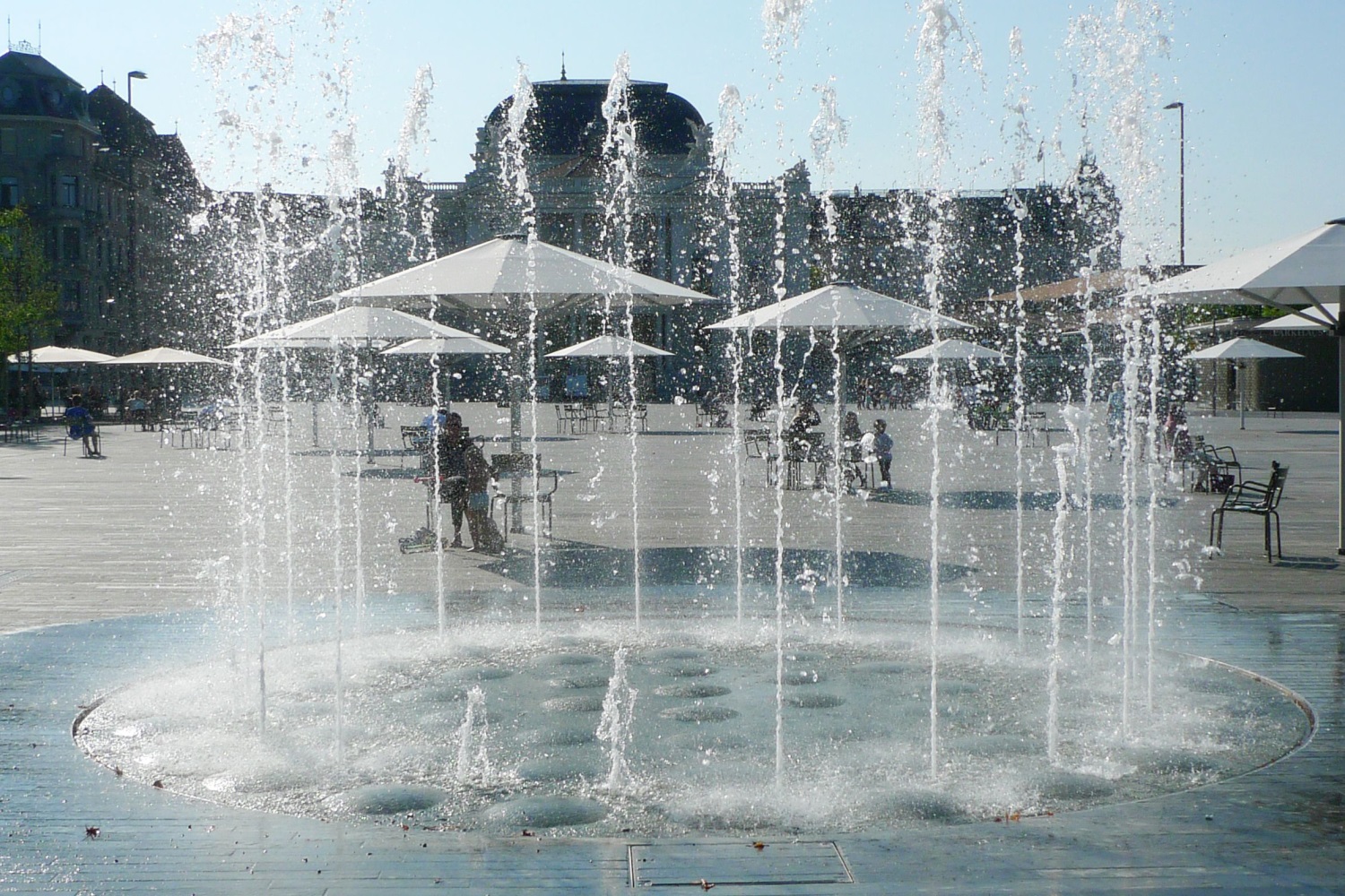 Das Bild zeigt das Wasserspiel und im Hintergrund Sonnenschirme und besetzte Stühle auf dem Sechseläutenplatz.
