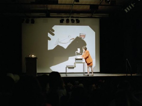 Frau in orangem Deux-pièce zeichnet auf Hellraumprojektor, Projektion ist hinter ihr zu sehen