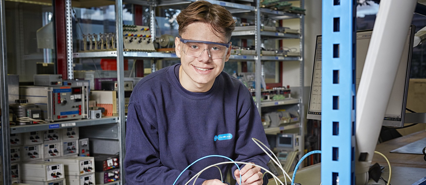 Ein/e lernende/r Automatiker/in EFZ mit dunkelblauem Pulli und Brille arbeitet in der Werkstatt mit Kabeln.