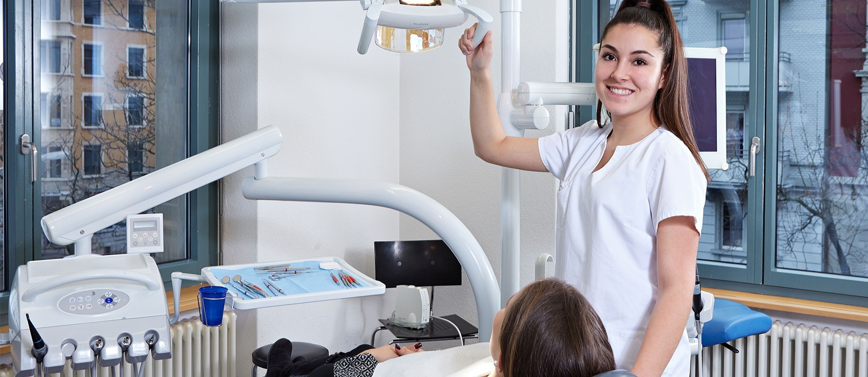 Ein/e lernende/r Dentalassistent/in EFZ in weisser Kleidung schiebt die Beleuchtung über den Kopf einer Kundin im Zahnarztstuhl.