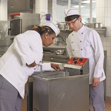 Eine Köchin erkärt einem fremdsprachigen Mitarbeiter die Fritteuse.