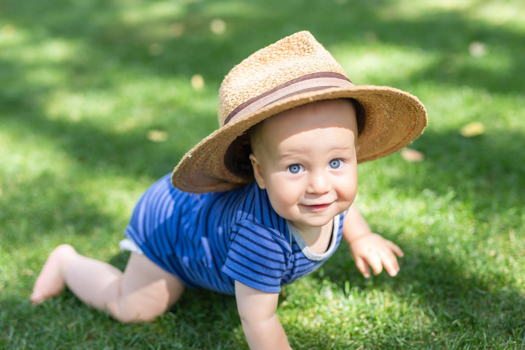 Portrait cute funny little boy in big straw hat having fun on green grass lawn at park. Sweet little baby crawling and smiling at garden on bright summer day. Happy childhood. Child activity outdoor