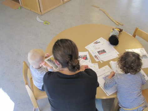 zwei Kinder an einem runden Tisch mit vielen beschrifteten Papieren drauf. Eine Person sitzt zwischen den Kindern und schaut die Blätter mit ihnen an. 