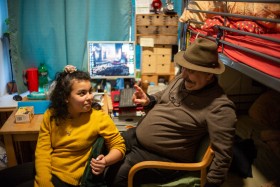 Bild eines Zimmer in der Notunterkunft für Familien in dem ein Vater im Zimmer sitzt Computer im Hintergrund und mit seiner Tochter spricht. Beide lächeln.