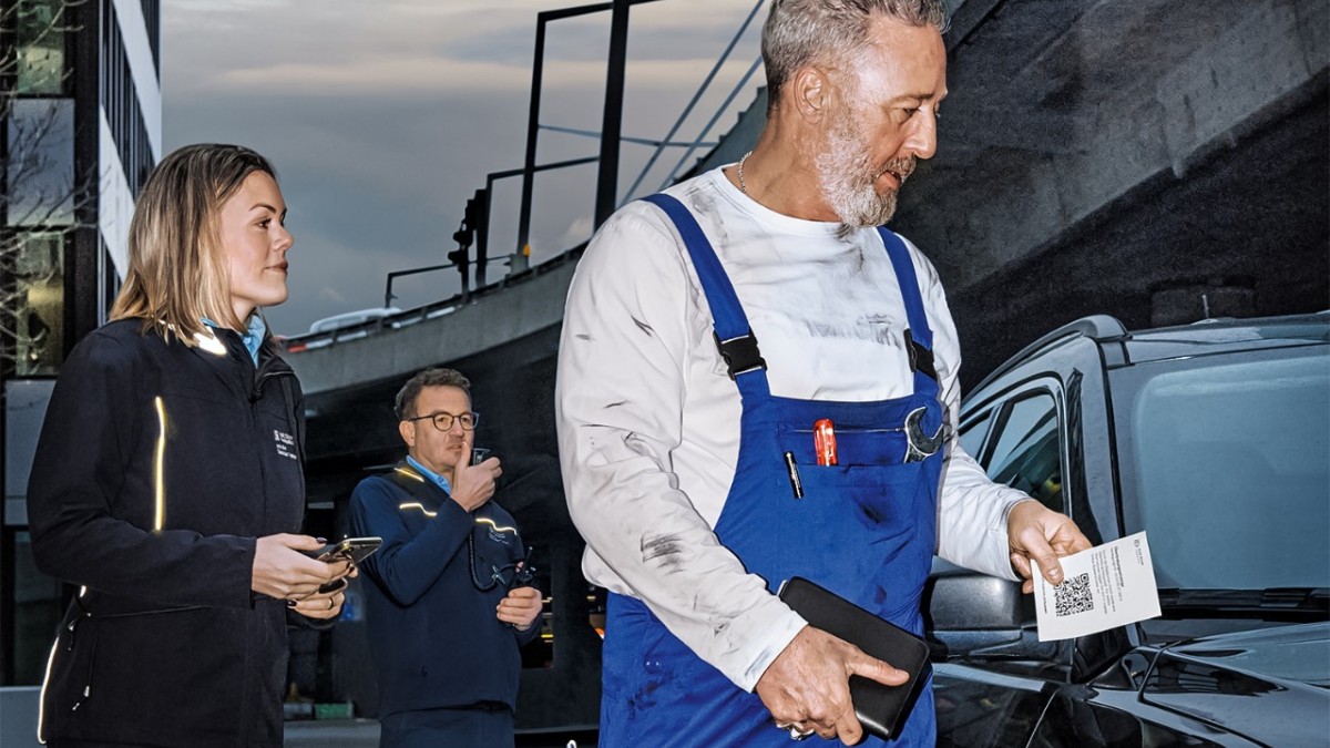 Ein Automechaniker mit einer Busse in der Hand. Hinter ihm zwei Polizist*innen, von denen einer in sein Funkgerät spricht.