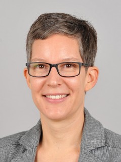 Porträt von Sabrina Wilhelmer: Eine lächelnde Frau mit ganz kurzen, dunkelgrauen Haaren und einer rechteckigen Brille
