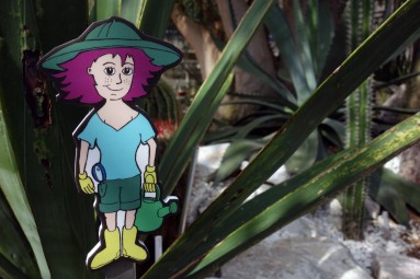 Die kleine Comicfigur von Sara Dorn steht im Beet vor einer Pflanze