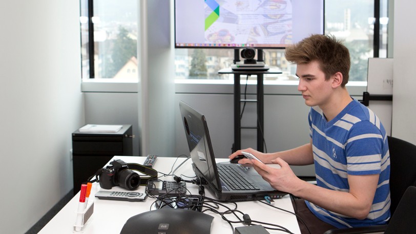 Junger Mann am Arbeitsplatz im Büro vor dem Notebook sitzend, in der Hand ein Smartphone.