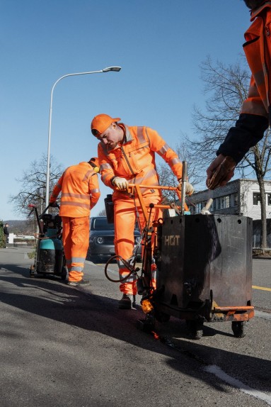 Drei Werkhofmitarbeiter vergiessen Risse auf einer Strasse mit einer grossen Maschine.