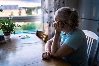 Ältere Frau sitzt an Tisch und sieht aus dem Fenster. Sie hat den Arm aufgestützt, sodass ihr Gesicht nicht erkennbar ist. Die Frau wirkt nachdenklich oder traurig. Ältere Frau sitzt an Tisch und sieht aus dem Fenster. Sie hat den Arm aufgestützt, sodass ihr Gesicht nicht erkennbar ist. Die Frau wirkt nachdenklich oder traurig.
