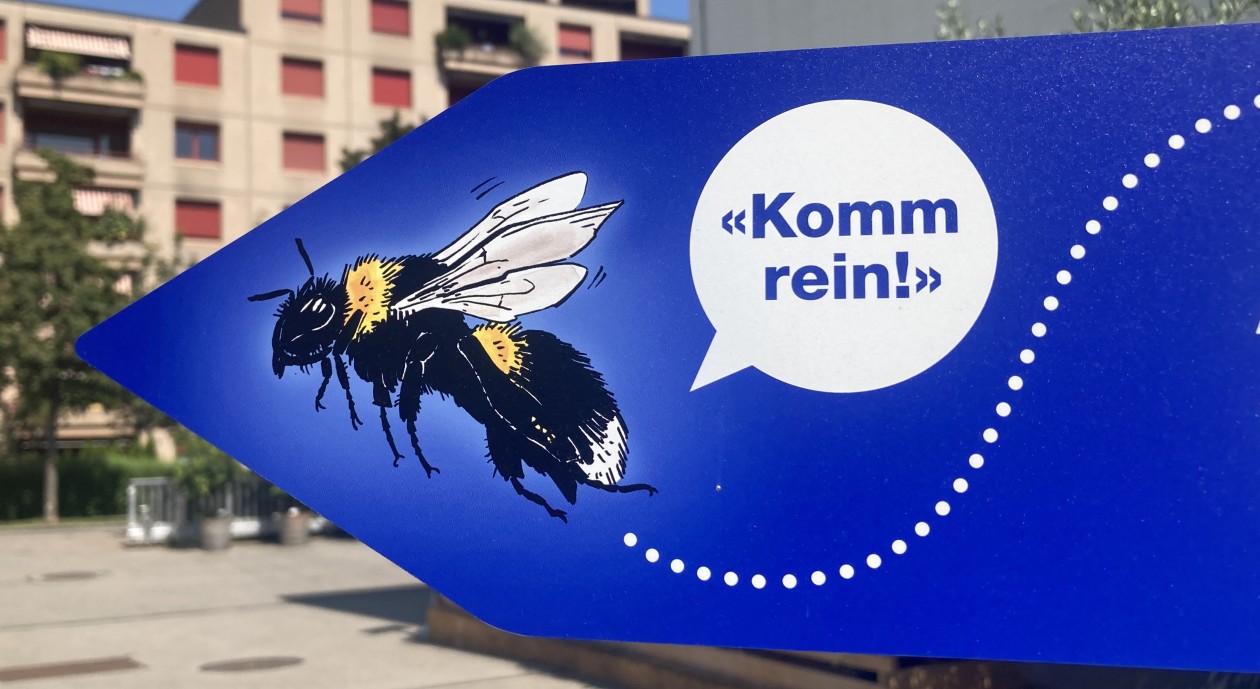 Ein Wegweiser-Schild auf dem Gelände der Stadtgärtnerei zeigt eine Hummel und eine Sprechblase mit dem Inhalt Komm Rein!