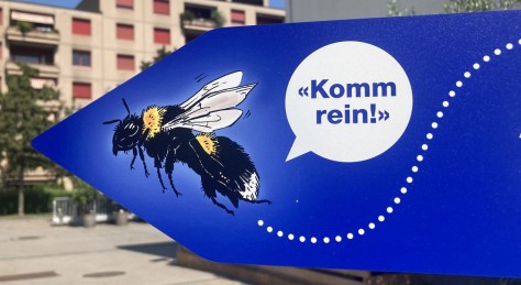 Ein Wegweiser-Schild auf dem Gelände der Stadtgärtnerei zeigt eine Hummel und eine Sprechblase mit dem Inhalt Komm Rein!
