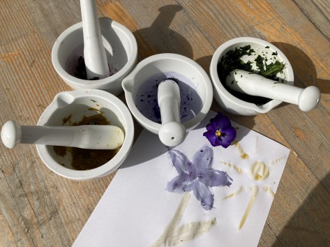 Auf einem Tisch stehen mehrere Mörser mit verschiedenenNaturmaterialiene. Daneben liegt ein selbstgemaltes Kinderbild., dass eine Blume und eine Sonne zeigt.